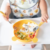 A baby in a high chair eats from a yellow plate, holding a fork and spoon while wearing a Bumkins SuperBib® 3 Pack: Pooh Bear and Friends.