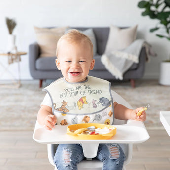 In a cozy living room, a toddler in a high chair wears a Bumkins SuperBib® 3 Pack: Pooh Bear and Friends while the tray holds a plate.
