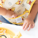 Close-up of a baby wearing a Bumkins SuperBib® 3 Pack: Pooh Bear and Friends, reaching into the front pocket.