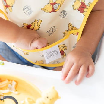 Close-up of a baby wearing a Bumkins SuperBib® 3 Pack: Pooh Bear and Friends, reaching into the front pocket.