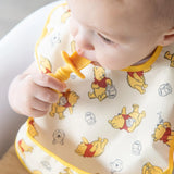 Close-up of a baby wearing a yellow Bumkins SuperBib® 3 Pack: Pooh Bear and Friends, chewing a yellow spoon.