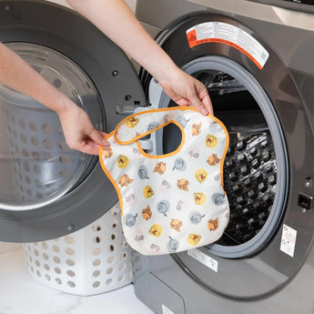 A person holds a Bumkins SuperBib® 3 Pack: Pooh Bear and Friends in front of a front-loading washing machine.