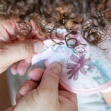 Hands fastening a Bumkins Love You Bunches SuperBib® 3 Pack with adjustable fit around a curly-haired childs neck.