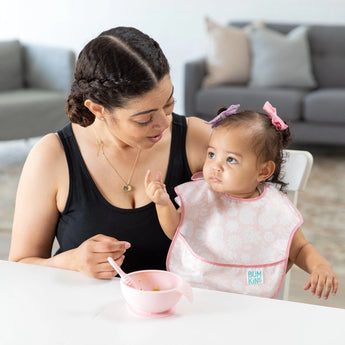 A woman and toddler with pink bows sit at a table; the toddler uses a bowl and spoon wearing Bumkins adjustable SuperBib® from Love You Bunches.