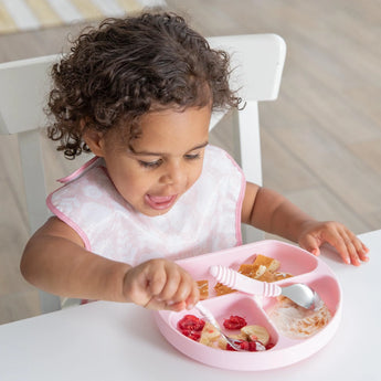 A curly-haired child enjoys fruit and yogurt on a pink plate, wearing a waterproof, adjustable SuperBib® from Bumkins.
