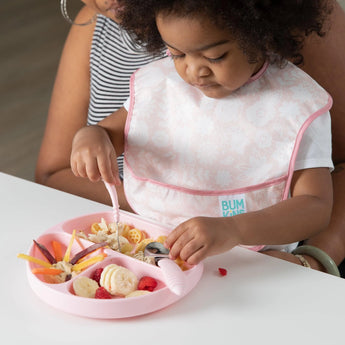 A toddler in a SuperBib® by Bumkins eats from a pink plate with a fork and spoon, assisted by an adults hand.