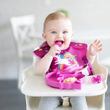 A happy baby in a high chair enjoys meal time with Bumkins Silicone Chewtensils® in Fuchsia and a pink plate, wearing a unicorn bib.