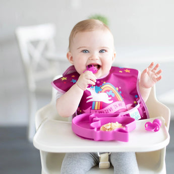 A happy baby in a high chair enjoys meal time with Bumkins Silicone Chewtensils® in Fuchsia and a pink plate, wearing a unicorn bib.
