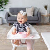 A smiling baby in a high chair with a pink bowl and spoon wears a Bumkins SuperBib® and Sleeved Bib 3-Piece Set: Always Hungry, XOXO, Hearts.