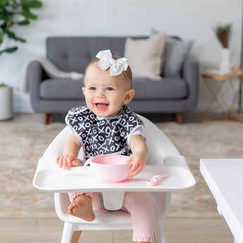 A smiling baby in a high chair with a pink bowl and spoon wears a Bumkins SuperBib® and Sleeved Bib 3-Piece Set: Always Hungry, XOXO, Hearts.