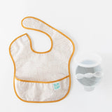 A beige Bumkins SuperBib® 3 Pack: Desert Boho lays flat next to a grey bowl and spoon on a white background.