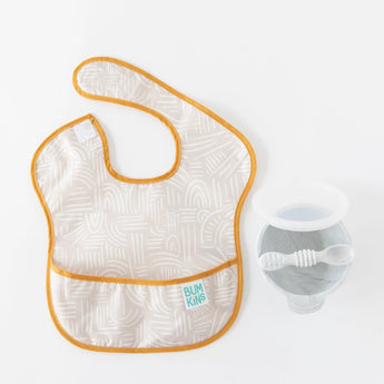 A beige Bumkins SuperBib® 3 Pack: Desert Boho lays flat next to a grey bowl and spoon on a white background.