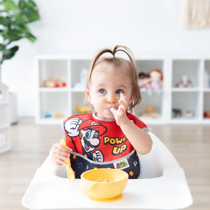 A toddler in a high chair wearing a Bumkins SuperBib® 3 Pack: Super Mario™ Power-Up eats from a yellow bowl.