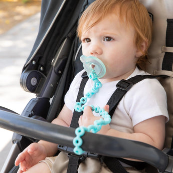 A baby with light brown hair sits in a stroller, with a pacifier attached to a Bumkins Silicone Accessory Tether 2-Pack: Blue.