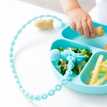 Close-up of a child reaching for food on a blue plate, with a toddler fork attached to a Bumkins Silicone Accessory Tether 2-Pack: Blue.
