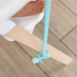Close-up of a baby's foot in a high chair, with a spoon hanging from a Bumkins Silicone Accessory Tether 2-Pack: Blue above the wood floor.