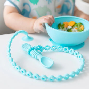 A baby in a patterned bib eats from a blue bowl at a table, with a blue spoon held by a Bumkins Silicone Accessory Tether 2-Pack: Blue.