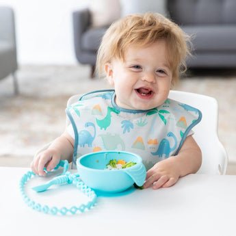 A happy baby in a dinosaur bib eats from a blue bowl at a table, with a spoon attached to a Bumkins Silicone Accessory Tether 2-Pack: Blue.
