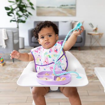 A toddler in a high chair wears a colorful bib, holding a spoon attached to the tray with a Bumkins Silicone Accessory Tether 2-Pack: Blue.