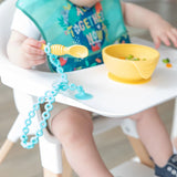 A baby explores a yellow bowl of food on a high chair tray, holding a spoon attached with a Bumkins Silicone Accessory Tether 2-Pack: Blue.