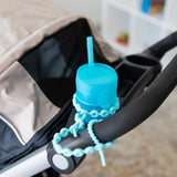 A blue cup with a straw in the cup holder of a stroller is secured to the handle with a Bumkins Silicone Accessory Tether 2-Pack: Blue.
