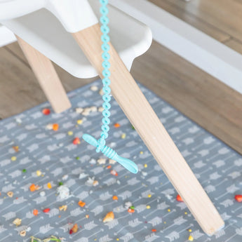 A spoon secured by a Bumkins Silicone Accessory Tether 2-Pack: Blue dangles from a high chair, as food rests on a patterned mat below.