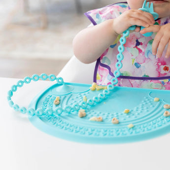 A child in a bib chews a silicone fork attached to a blue placemat with a Bumkins Silicone Accessory Tether 2-Pack: Blue.