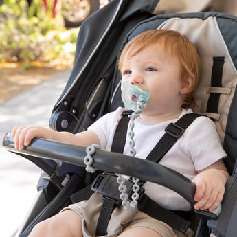 A baby in a stroller outdoors using a pacifier attached to a Bumkins Silicone Accessory Tether 2-Pack: Gray, with greenery behind.