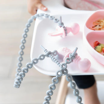 Close-up of pink toddler utensils attached to Bumkins Silicone Accessory Tether 2-Pack: Gray on a high chair tray.