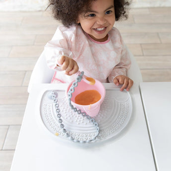 A toddler eats from a pink bowl on a gray placemat, holding a spoon attached to a Bumkins Silicone Accessory Tether 2-Pack: Gray.