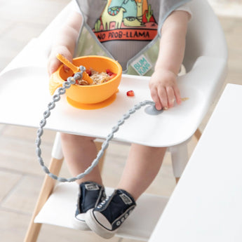 A baby in a bib and sneakers eats from a bowl in a high chair, with a spoon attached to a Bumkins Silicone Accessory Tether 2-Pack: Gray.