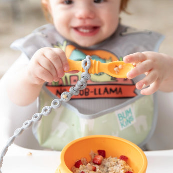 A child holds a spoon attached to a Bumkins Silicone Accessory Tether 2-Pack: Gray over a bowl of oatmeal on a high chair tray.
