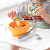 A baby in a high chair eats from an orange bowl, with a spoon attached to the tray with a Bumkins Silicone Accessory Tether 2-Pack: Gray.