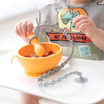 A baby in a high chair eats from an orange bowl, with a spoon attached to the tray with a Bumkins Silicone Accessory Tether 2-Pack: Gray.