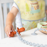 Close-up of a child in a colorful bib at a table, holding a toddler utensil secured by a Bumkins Silicone Accessory Tether 2-Pack: Gray.