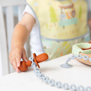Close-up of a child in a colorful bib at a table, holding a toddler utensil secured by a Bumkins Silicone Accessory Tether 2-Pack: Gray.