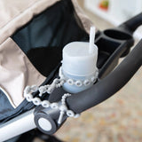 A gray cup in a stroller's cup holder is secured to the handle by a Bumkins Silicone Accessory Tether 2-Pack: Gray.