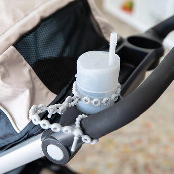A gray cup in a stroller's cup holder is secured to the handle by a Bumkins Silicone Accessory Tether 2-Pack: Gray.