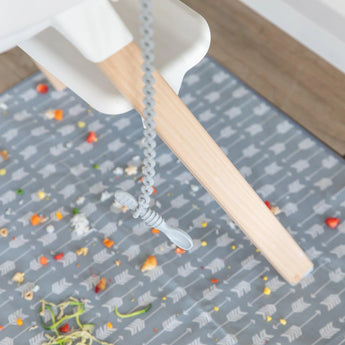 A spoon secured by a Bumkins Silicone Accessory Tether 2-Pack: Gray dangles from a high chair, as food rests on a patterned mat below.