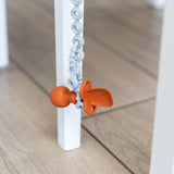 Close-up of an orange toddler spoon hanging from a Bumkins Silicone Accessory Tether 2-Pack: Gray next to a white chair leg on a wood floor.
