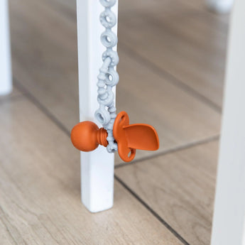 Close-up of an orange toddler spoon hanging from a Bumkins Silicone Accessory Tether 2-Pack: Gray next to a white chair leg on a wood floor.