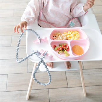 A baby in a high chair enjoys food from a plate, with toddler utensils secured to the tray by Bumkins Silicone Accessory Tether 2-Pack: Gray.