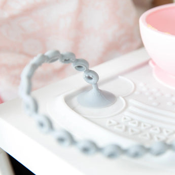 Close-up of a Bumkins Silicone Accessory Tether 2-Pack: Gray on a high chair tray, with the suction cup attached to a placemat.