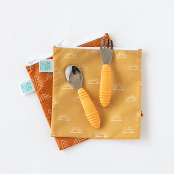 Bumkins Reusable Snack Bag, Large 2-Pack: Sunshine and Grounded lay overlapped on a white background, with yellow utensils on top. 
