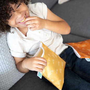 A child in a white shirt and jeans laughs on a couch, holding Bumkins Reusable Snack Bag, Large 2-Pack: Sunshine and Grounded.