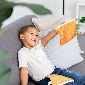 A child sits on a couch, smiling as he holds a Bumkins Reusable Snack Bag, Large 2-Pack: Sunshine and Grounded, with the other bag on his lap.