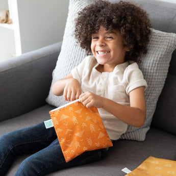 A curly-haired child smiles on a gray couch, holding an orange Bumkins Reusable Snack Bag, Large 2-Pack: Sunshine and Grounded.