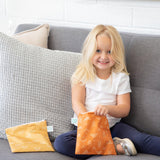 A smiling girl on a sofa reaches into a Bumkins Reusable Snack Bag, Large 2-Pack: Sunshine and Grounded, with the other bag nearby.