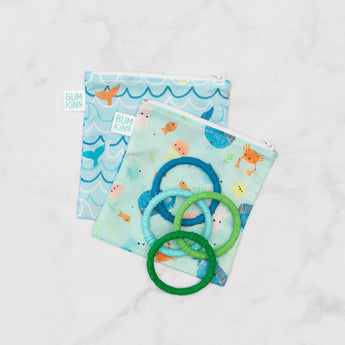 The Bumkins Reusable Snack Bag 2-Pack in Ocean Life & Whale Tail prints securely holds silicone teething rings on white marble.