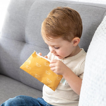 A child on a gray couch peers into a yellow Bumkins Reusable Snack Bag, Small 2-Pack: Sunshine and Grounded.